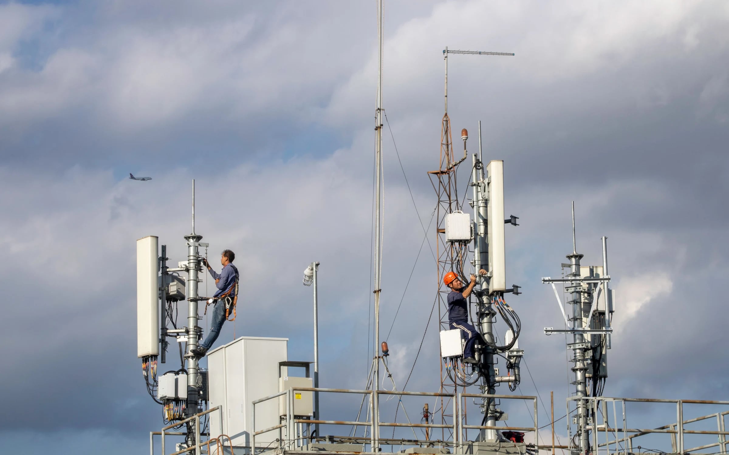 Technicians working on a cellular antenna tower under a cloudy sky