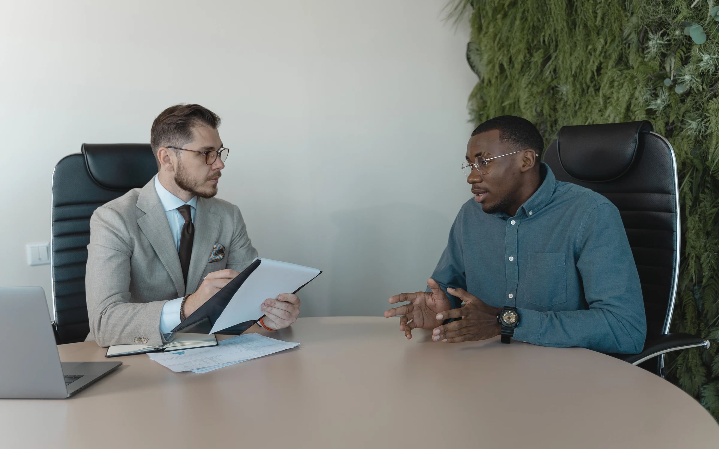 Two people talking across a round table in a meeting room, one taking notes
