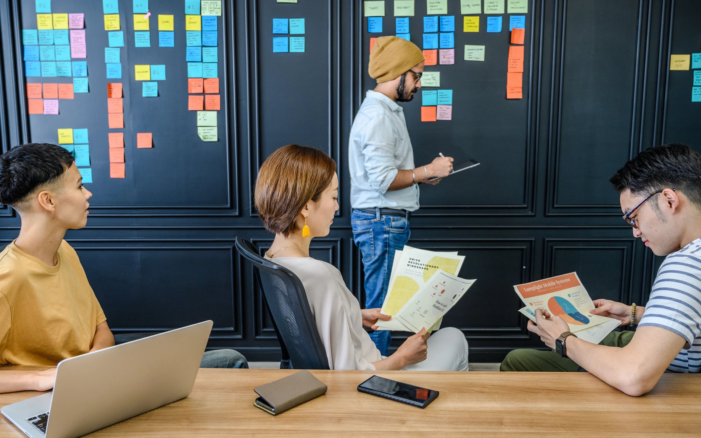 A team reviewing printed journey maps beside a wall of sticky notes in a modern workspace